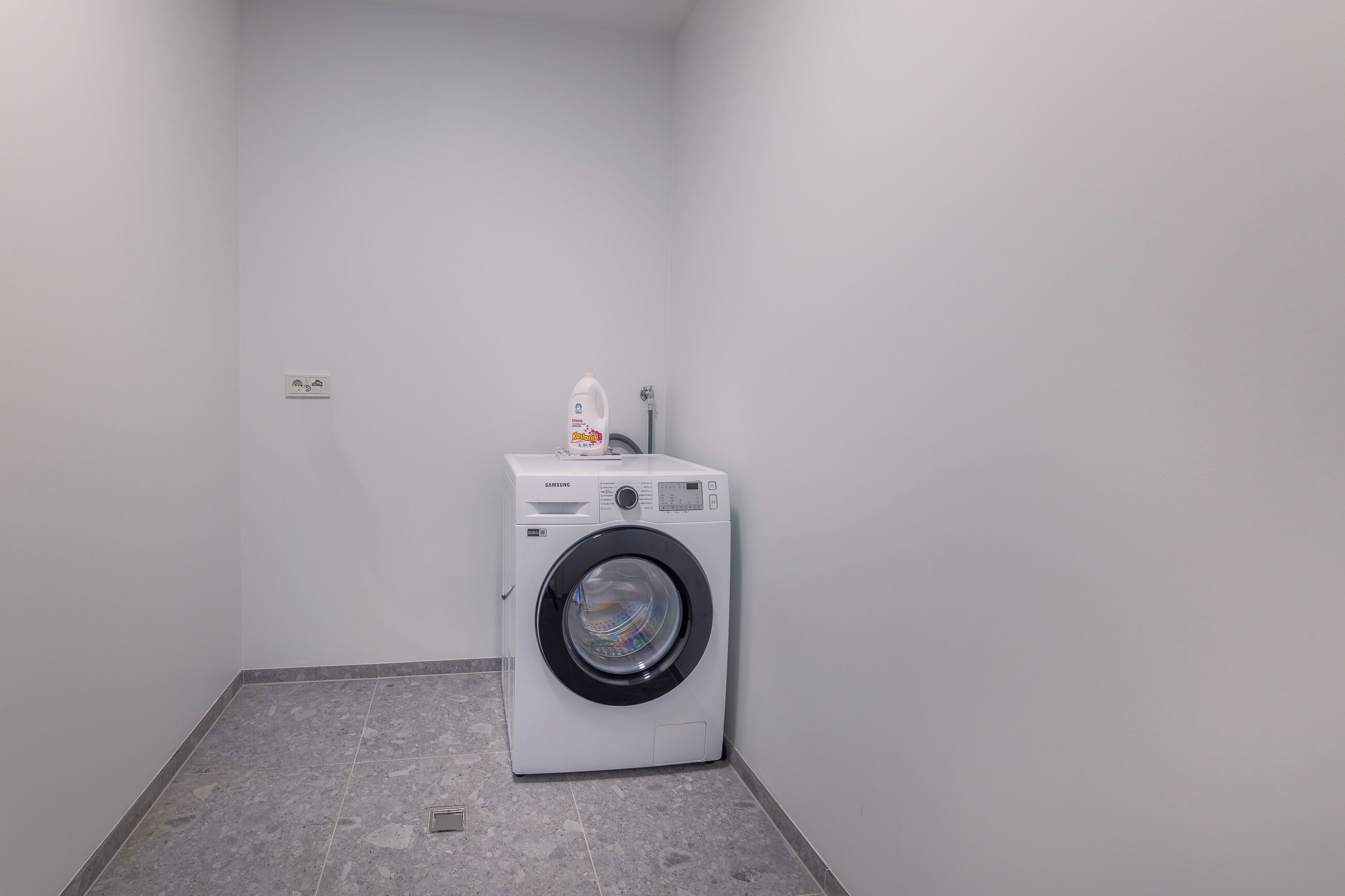 A small laundry room with a front-loading washing machine and a detergent bottle on top.