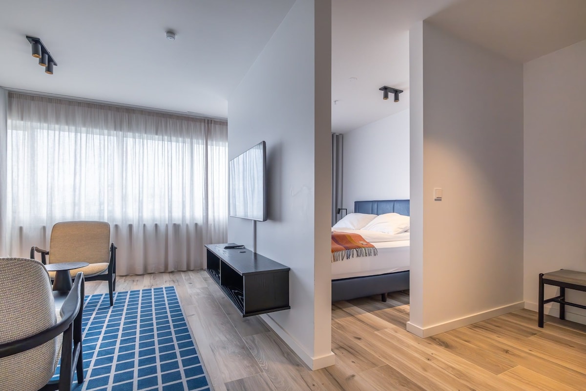 A modern hotel room with a bed, a desk, a chair, a TV, and a large window with curtains.
