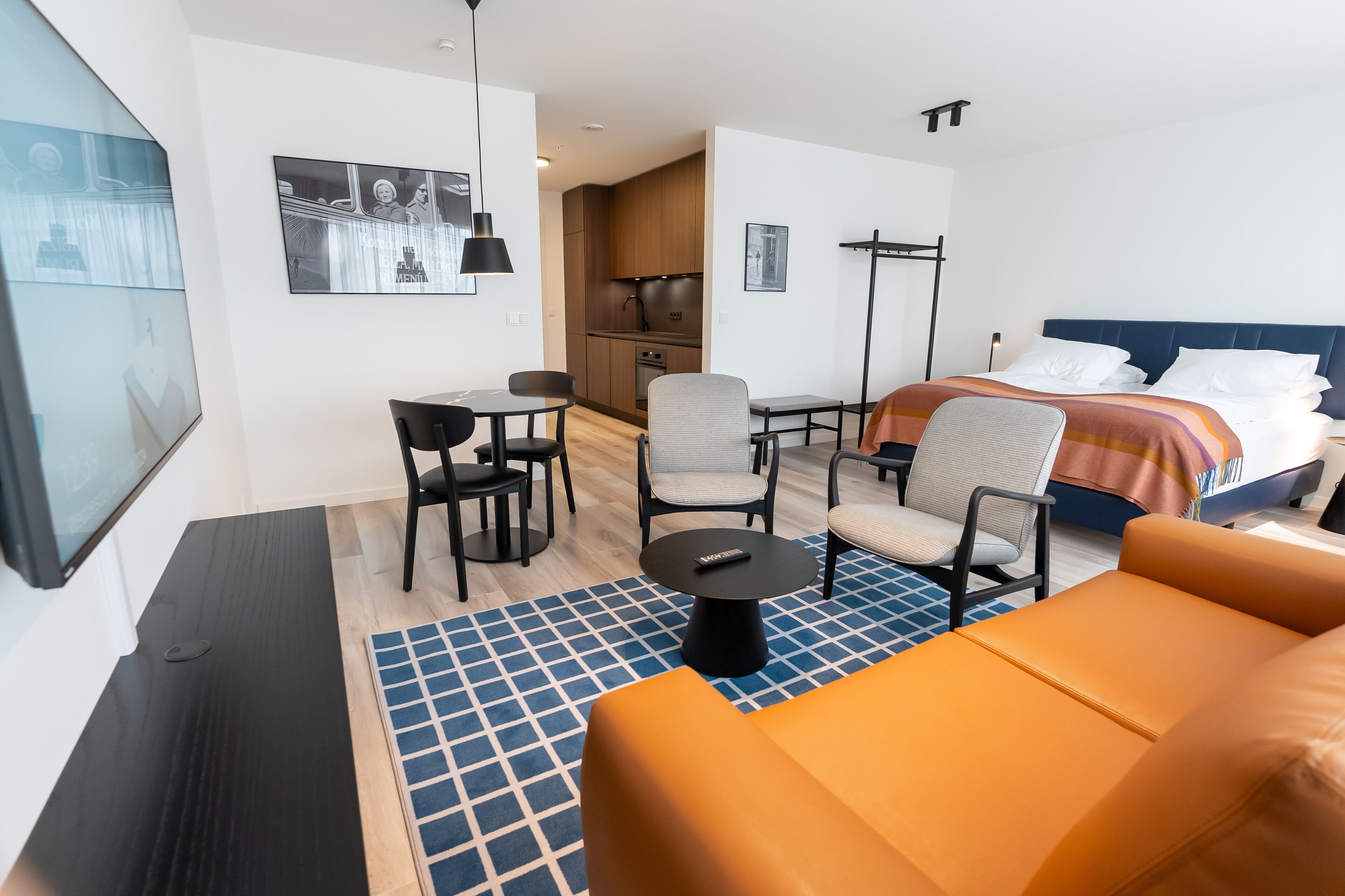 A modern hotel room with a bed, a desk, a chair, a sofa, and a small kitchen area.