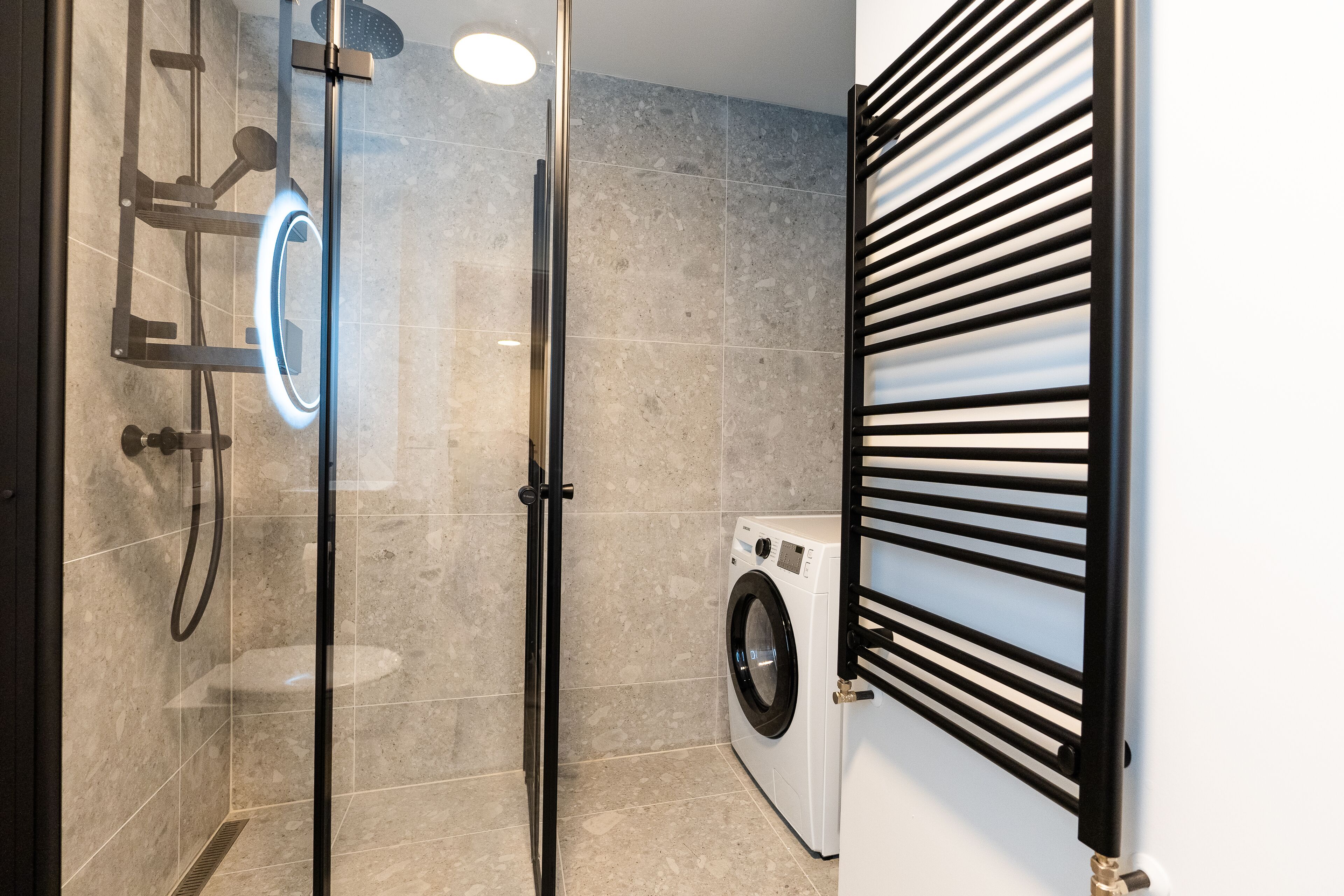 A modern bathroom with a glass-enclosed shower, a built-in washing machine, and a black metal rack.