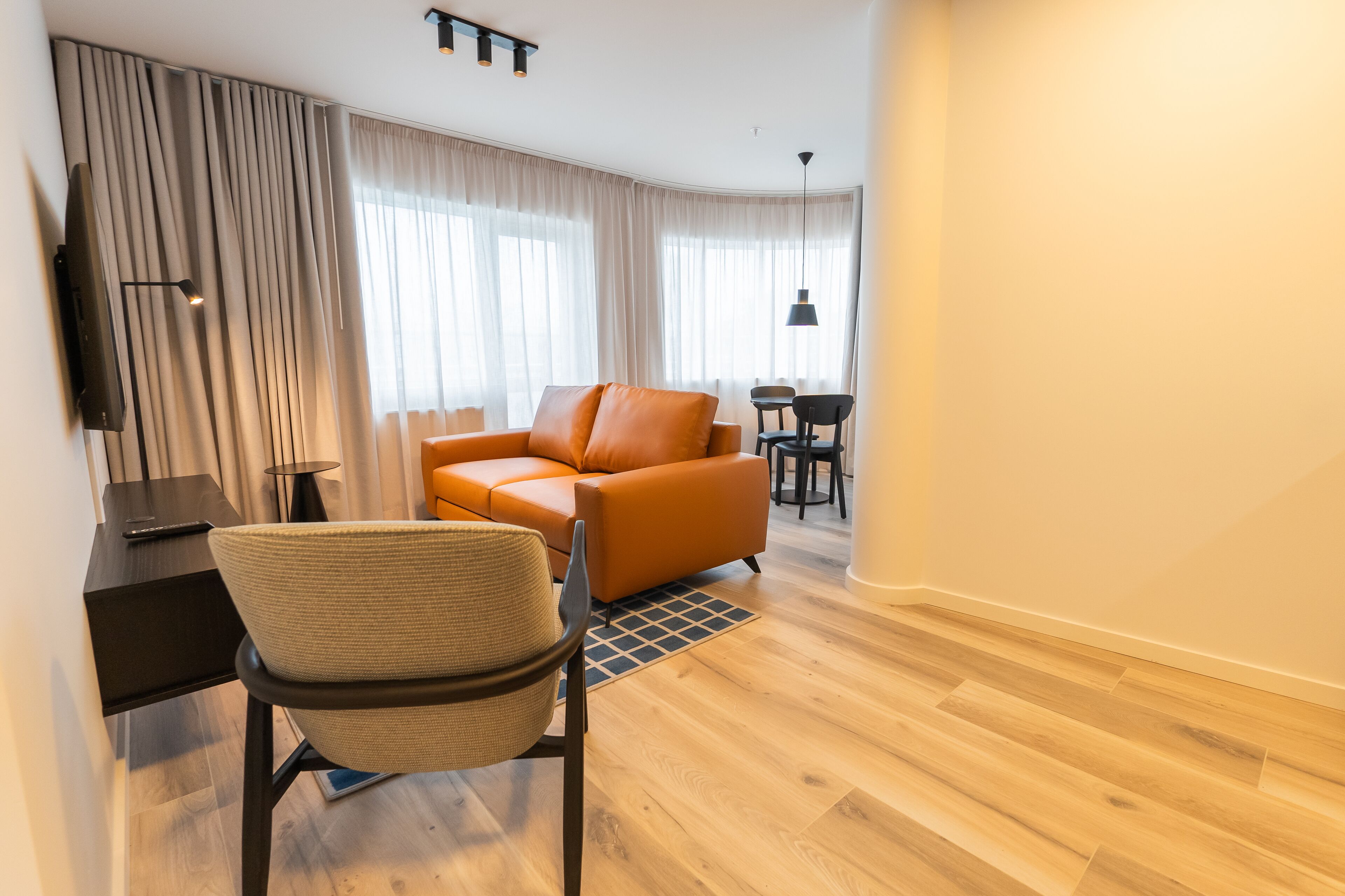 A modern living room with a leather sofa, a woven chair, a TV mounted on the wall, and a dining area with a table and chairs.
