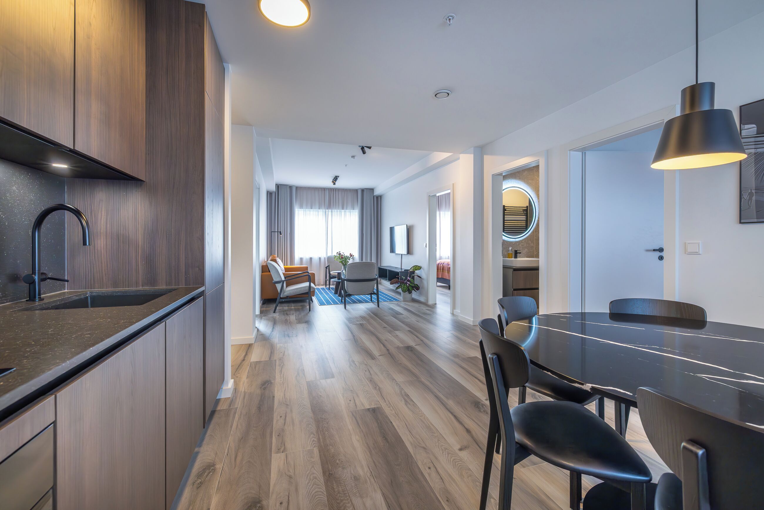 Une cuisine moderne avec des meubles en bois foncé et un plan de travail noir. On y trouve un coin repas avec une table et des chaises noires. La pièce est dotée d’un parquet et d’une grande fenêtre avec des rideaux.