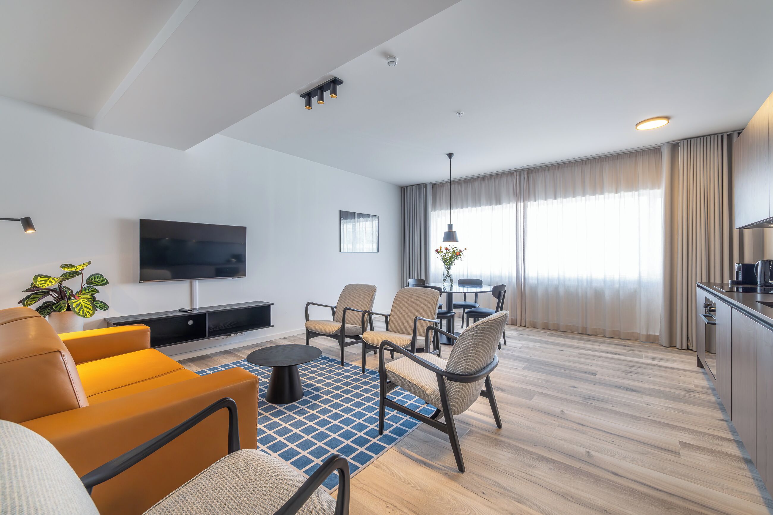 A modern living room with a flat-screen TV, comfortable seating, a small coffee table, and a kitchen area with a dining table.