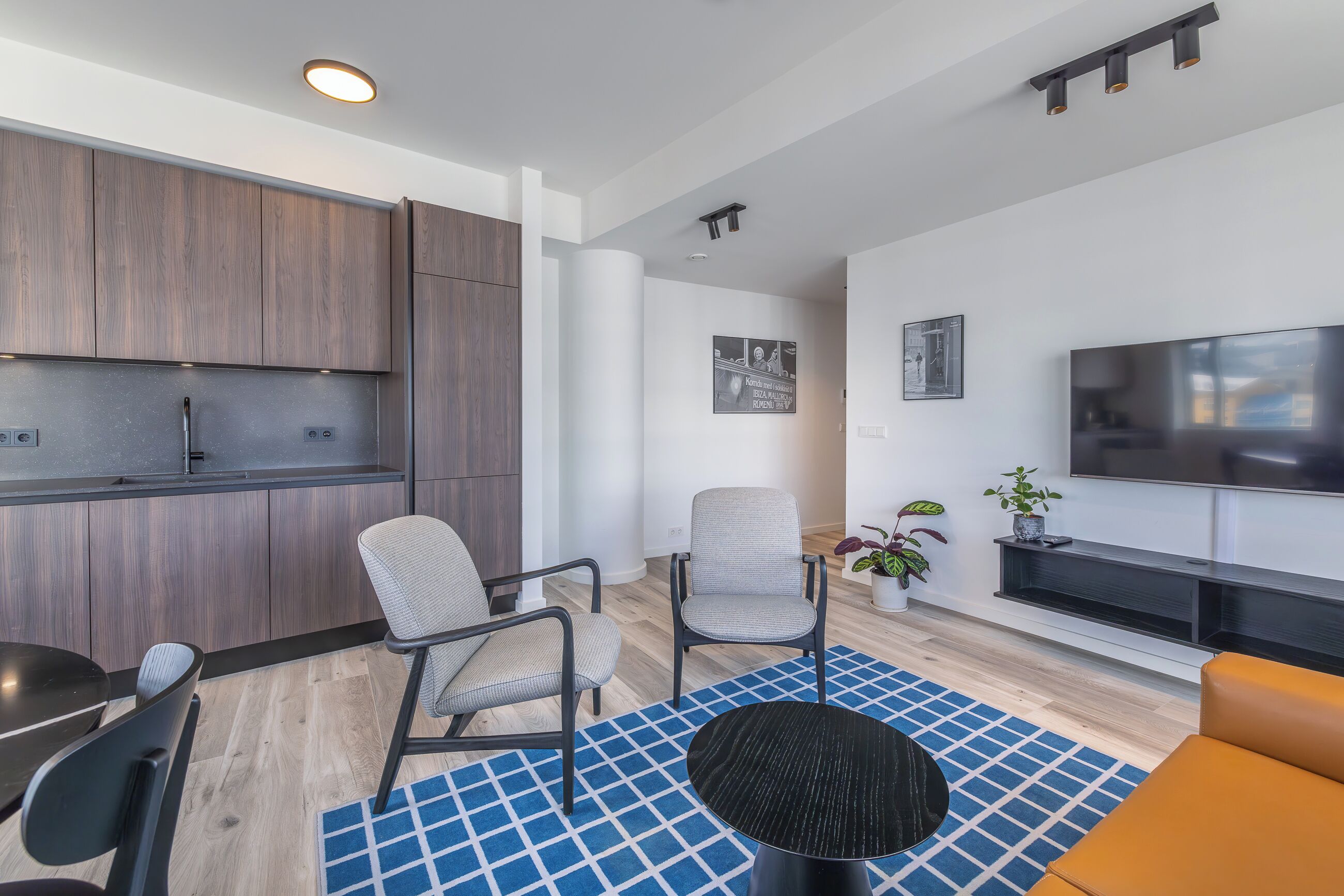 A modern living room with a flat-screen TV, two grey chairs, a black coffee table, and a blue patterned rug.