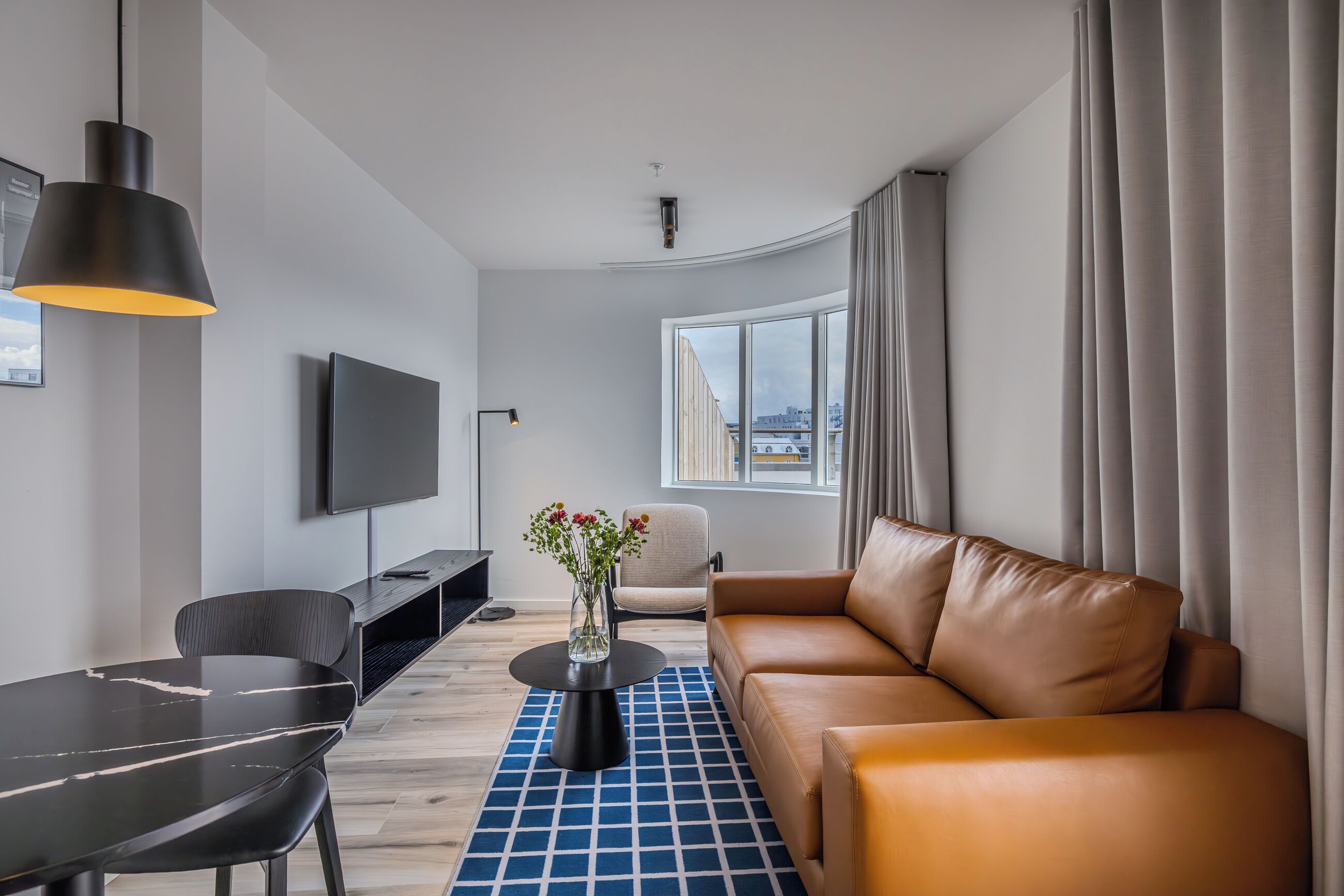 Una sala de estar moderna con un sofá de cuero, una mesa de comedor, un televisor y un amplio ventanal con cortinas.