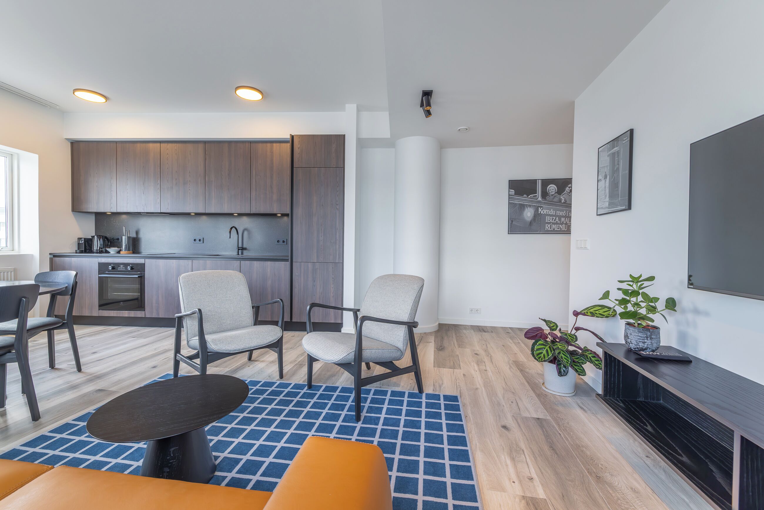 Una sala de estar moderna con una cocina al fondo, que presenta armarios de madera, un comedor con sillas, una mesa de centro y un televisor sobre un mueble.