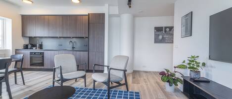 Una sala de estar moderna con una cocina al fondo, que presenta armarios de madera, un comedor con sillas, una mesa de centro y un televisor sobre un mueble.