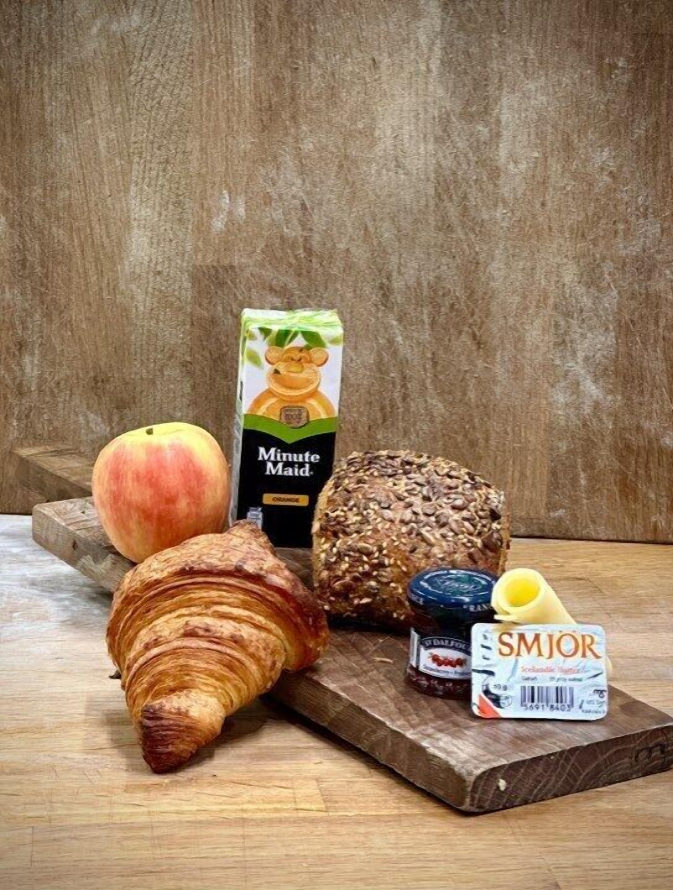 Een houten tafel met een croissant, een perzik, een brood, een pak Minute Maid sinaasappelsap, een pot jam en een pak SMJOR kaasverspreiding.