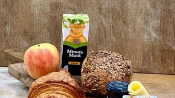 Een houten tafel met een croissant, een perzik, een brood, een pak Minute Maid sinaasappelsap, een pot jam en een pak SMJOR kaasverspreiding.