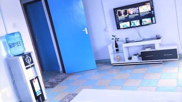 A room with a blue door, a white TV stand with a flat-screen TV, a white table, and a blue sofa. - Lux Suites Chief Engineer Apartments (Mwingi)