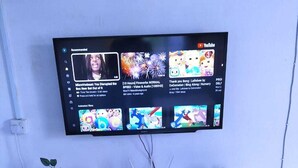 A flat-screen TV mounted on a wall above a white entertainment unit with a glass display case, a small plant, and a decorative item. - Lux Suites Chief Engineer Apartments (Mwingi)