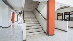 Un escalier aux marches blanches et aux piliers orange, menant à un étage supérieur dans un bâtiment dont le sol est recouvert de carreaux et les murs sont blancs.