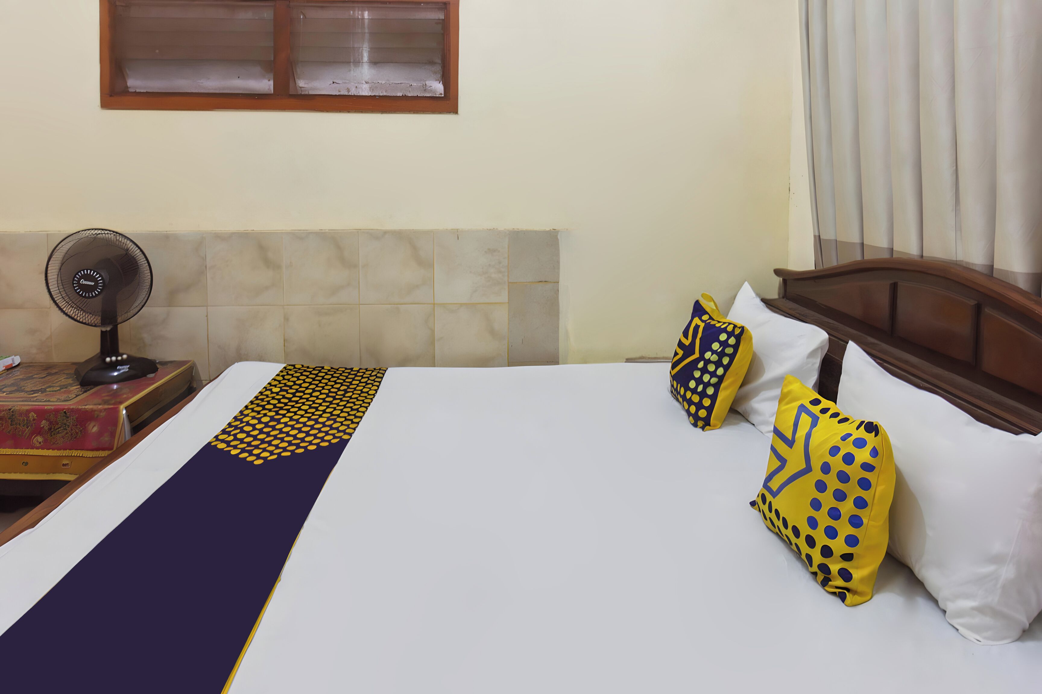 A hotel room with a bed, a standing fan, a window with blinds, and a curtain.