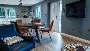 A modern dining area with a glass table, leather chairs, and a view of the outdoors through large windows. - Bramley Brook Cottage 5 star Luxury (Whalley)