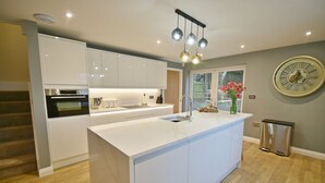 A modern kitchen with white cabinetry, a central island, a stainless steel sink, and a wall-mounted clock. - Bramley Brook Cottage 5 star Luxury (Whalley)