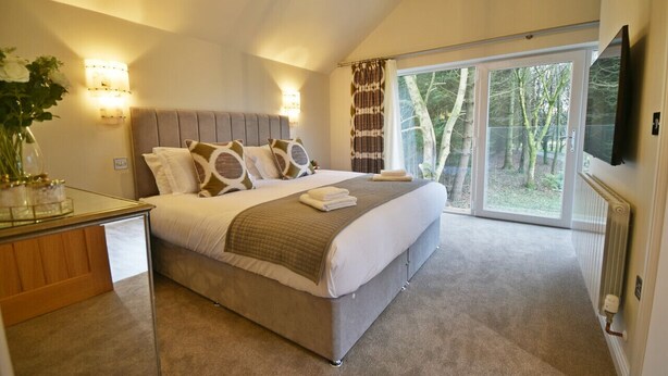 A bedroom with a large bed, two bedside lamps, a TV, and a view of trees through the window. - Bramley Brook Cottage 5 star Luxury (Whalley)