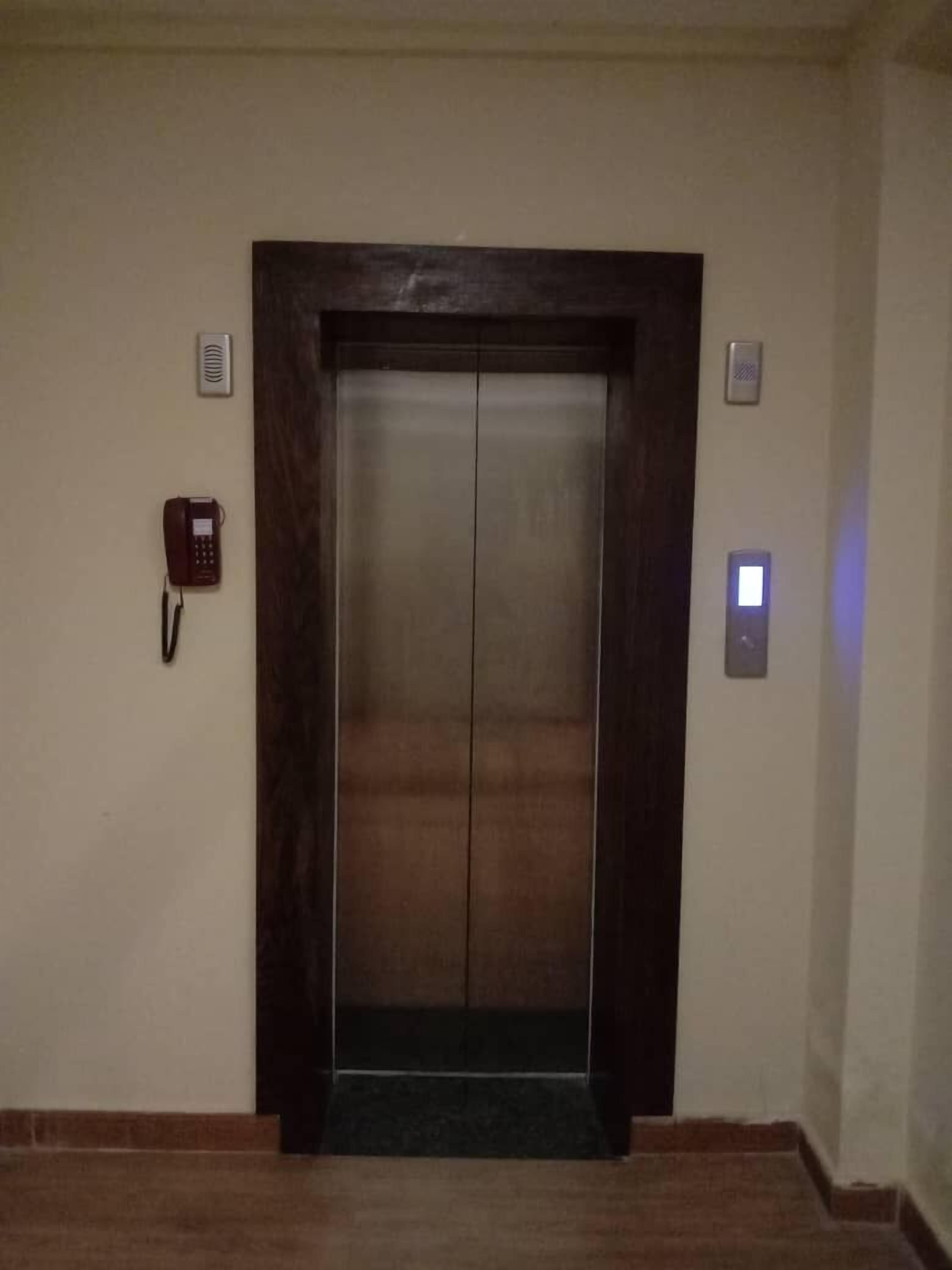 Un ascensor con estructura de madera oscura, un teléfono rojo en la pared a la izquierda y un interruptor de luz a la derecha.