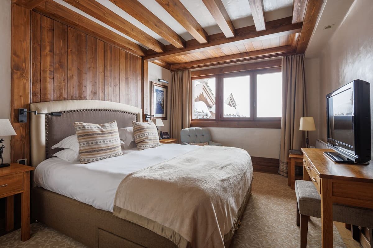 Quarto de hotel com uma cama grande, vigas de madeira no teto, televisão e janela com vista para um edifício.