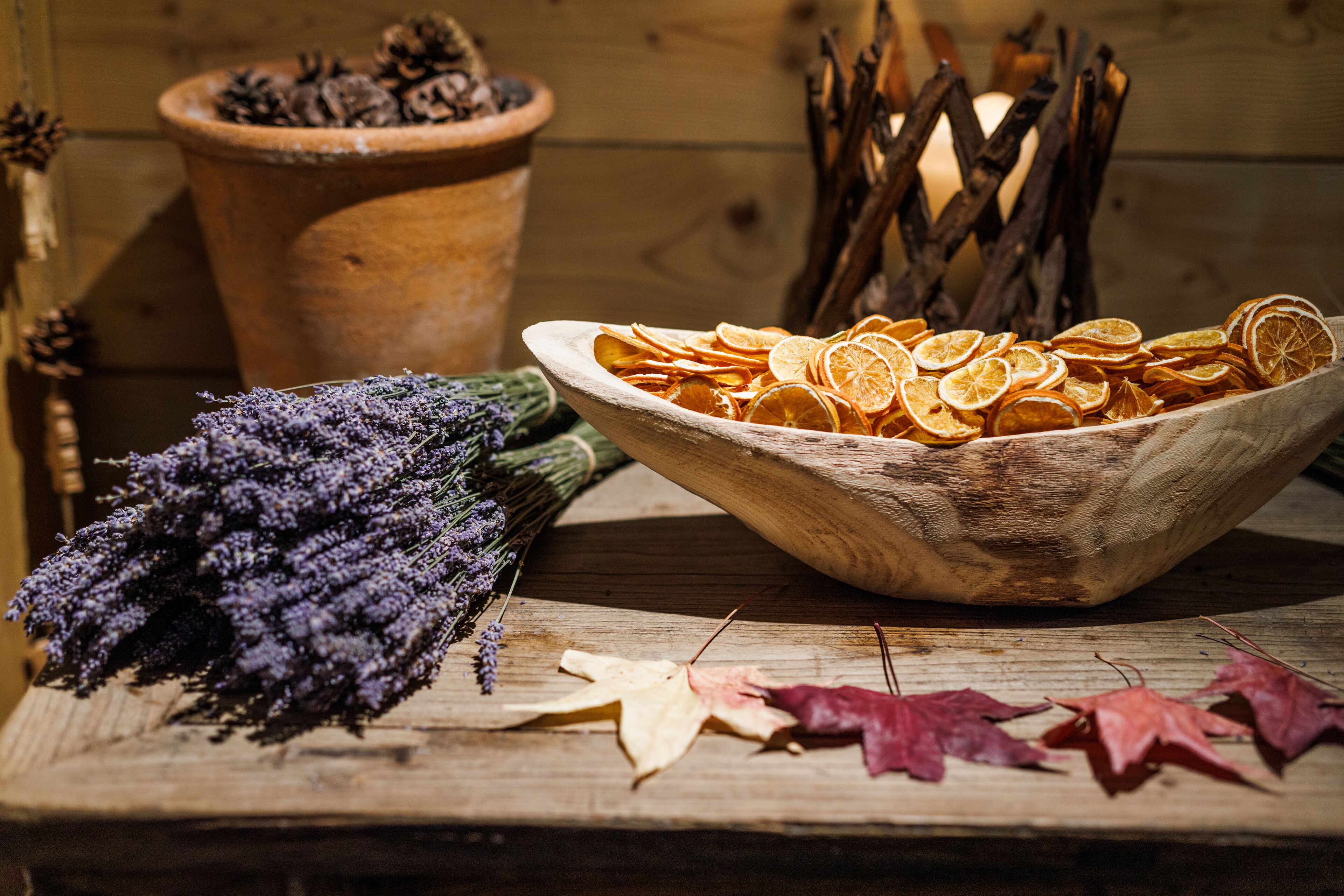 Une table en bois rustique, sur laquelle se trouve un bol rempli de tranches d’orange séchées, un bouquet de lavande et une pomme de pin dans un pot en terre cuite.