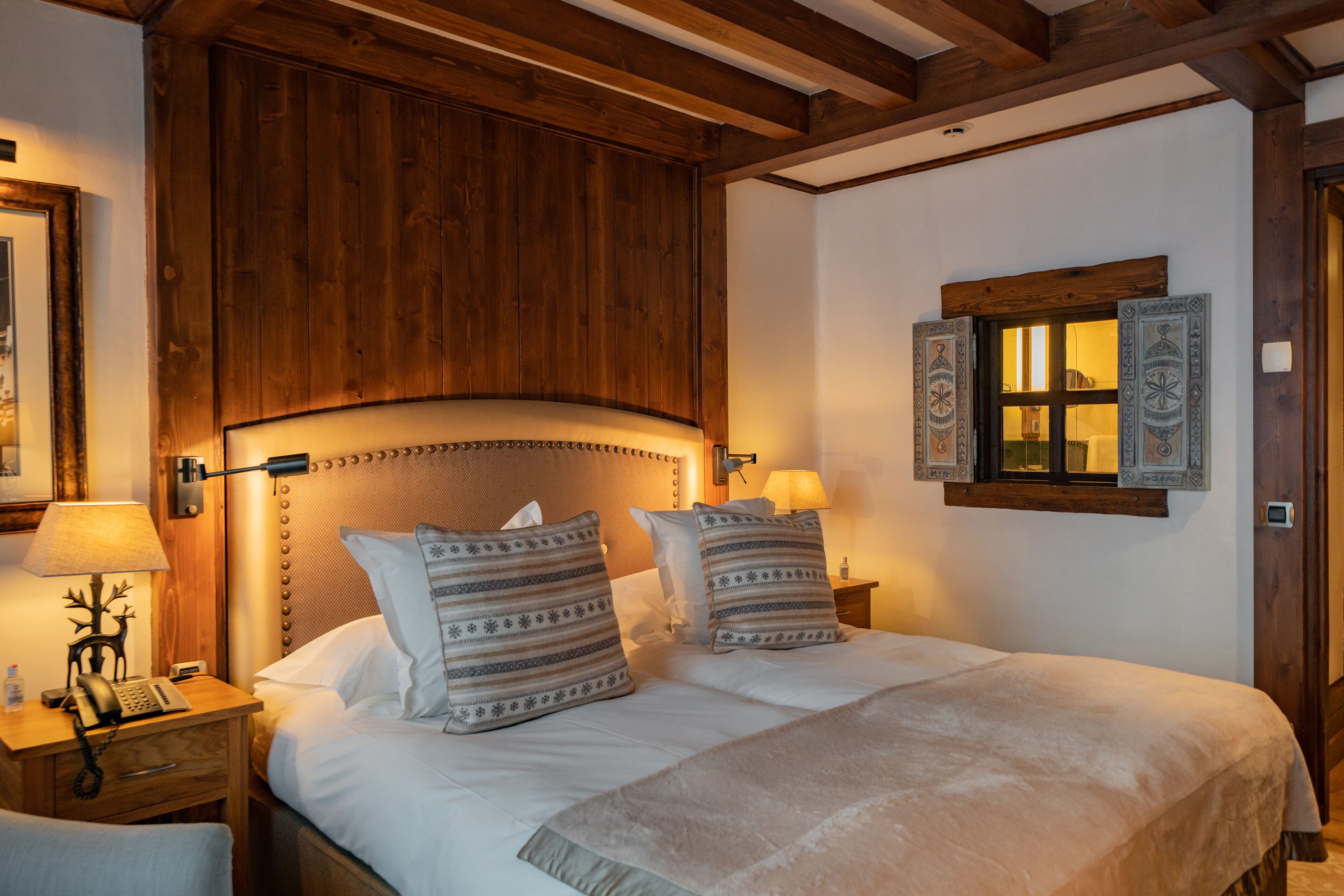 A hotel room with a wooden headboard, two bedside lamps, a bed with patterned pillows, a nightstand with a lamp, and a window with decorative shutters.