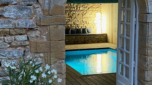 Un arc en pierre mène à un espace piscine agrémenté d’une terrasse en bois et d’une plante en pot.