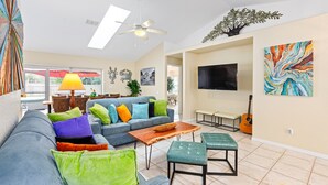 Ein Wohnzimmer mit Sofa, Couchtisch und Hocker, Deckenventilator, Fernseher und Bildern an den Wänden.
