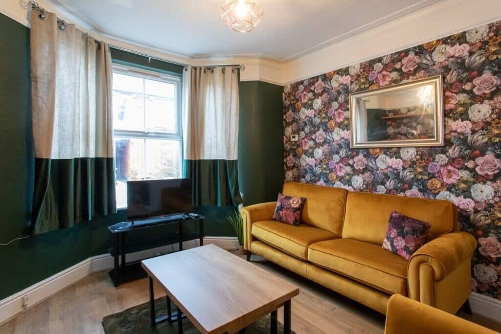 Um quarto com parede verde, sofá amarelo, mesa de centro de madeira e papel de parede floral.