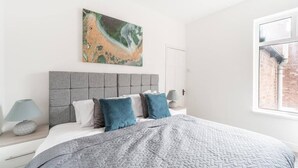 A modern bedroom with a grey headboard, a large abstract painting, and a view of a brick building through a window. - Near Hull Port House w/ Free Parking & Garden (Kingston upon Hull)