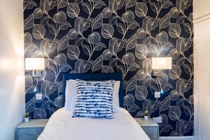Hotellihuone, jossa on sänky, kaksi yöpöytävalaisinta, tumma, lehtikuviollinen tapetti ja pieni kasvi yöpöydällä.