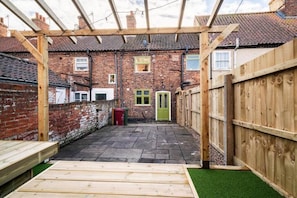 Un patio abrité avec une pergola en bois, un bâtiment en briques en arrière-plan et une porte verte.
