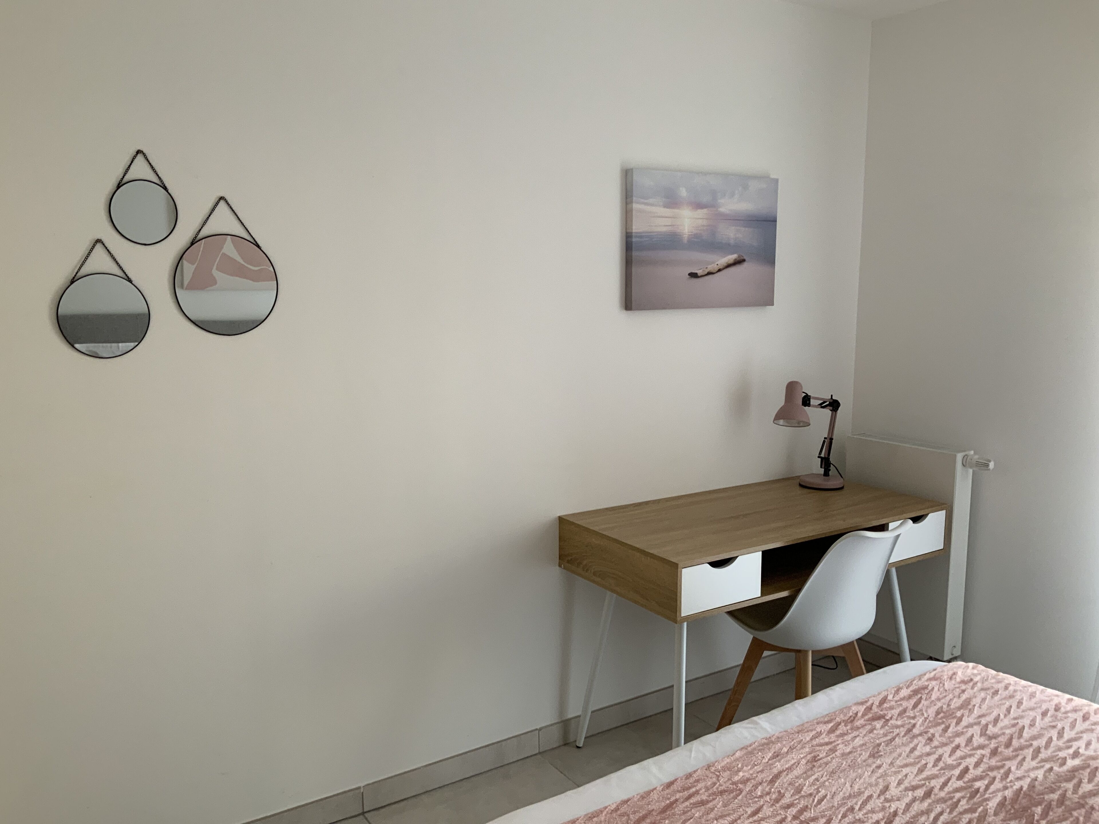Un dormitorio con un escritorio de madera, una silla, una cama con manta rosa y adornos en la pared que incluyen tres espejos y un cuadro.