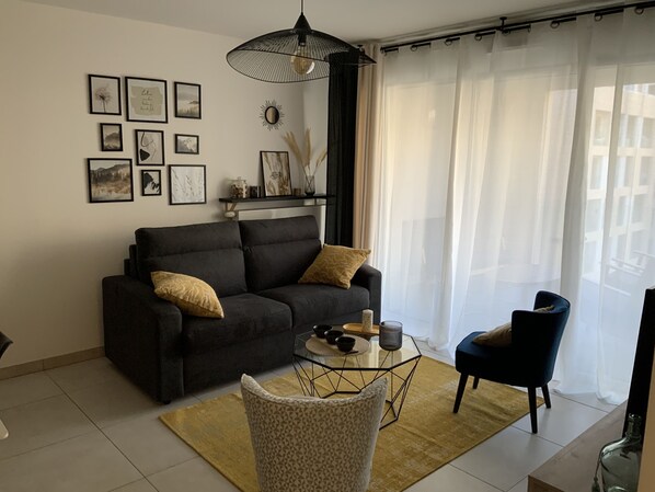 A living room with a dark sofa, a glass coffee table, and a blue armchair. There are framed pictures on the wall and a large window with sheer curtains. - Logement Cosy Avec Parking (Aix-en-Provence)