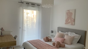 A bedroom with a bed, bedside tables, a desk, a chair, a window with curtains, and a ceiling light. - Logement Cosy Avec Parking (Aix-en-Provence)