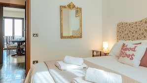 A hotel room with a bed, a large mirror, a bedside table with a lamp, and a view of the dining area. - Charming 4-bed Apartment -lake Views & Balconies (Oliveto Lario)