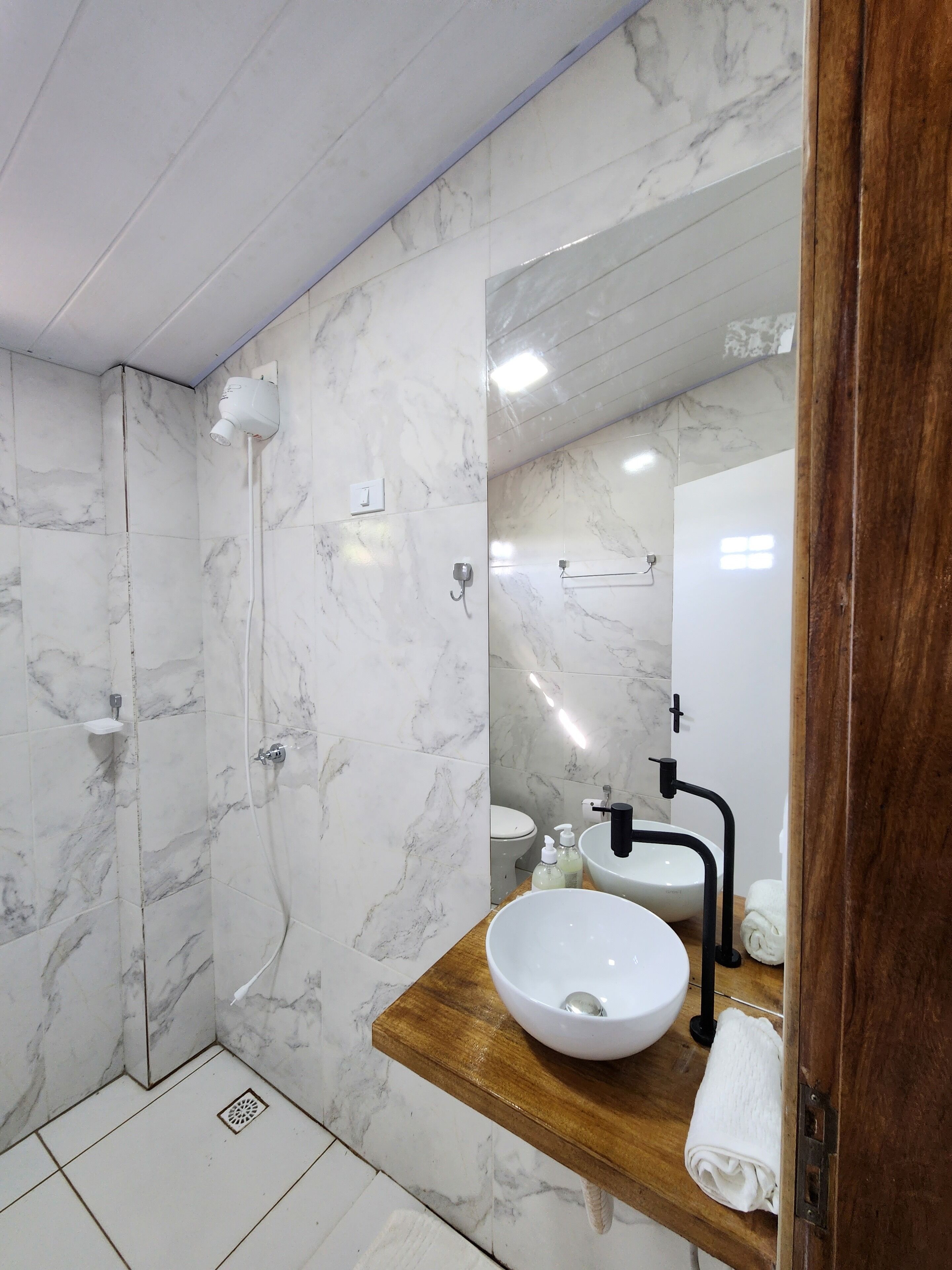 A modern bathroom with a marble wall, a wooden vanity, a white sink, and a shower area.