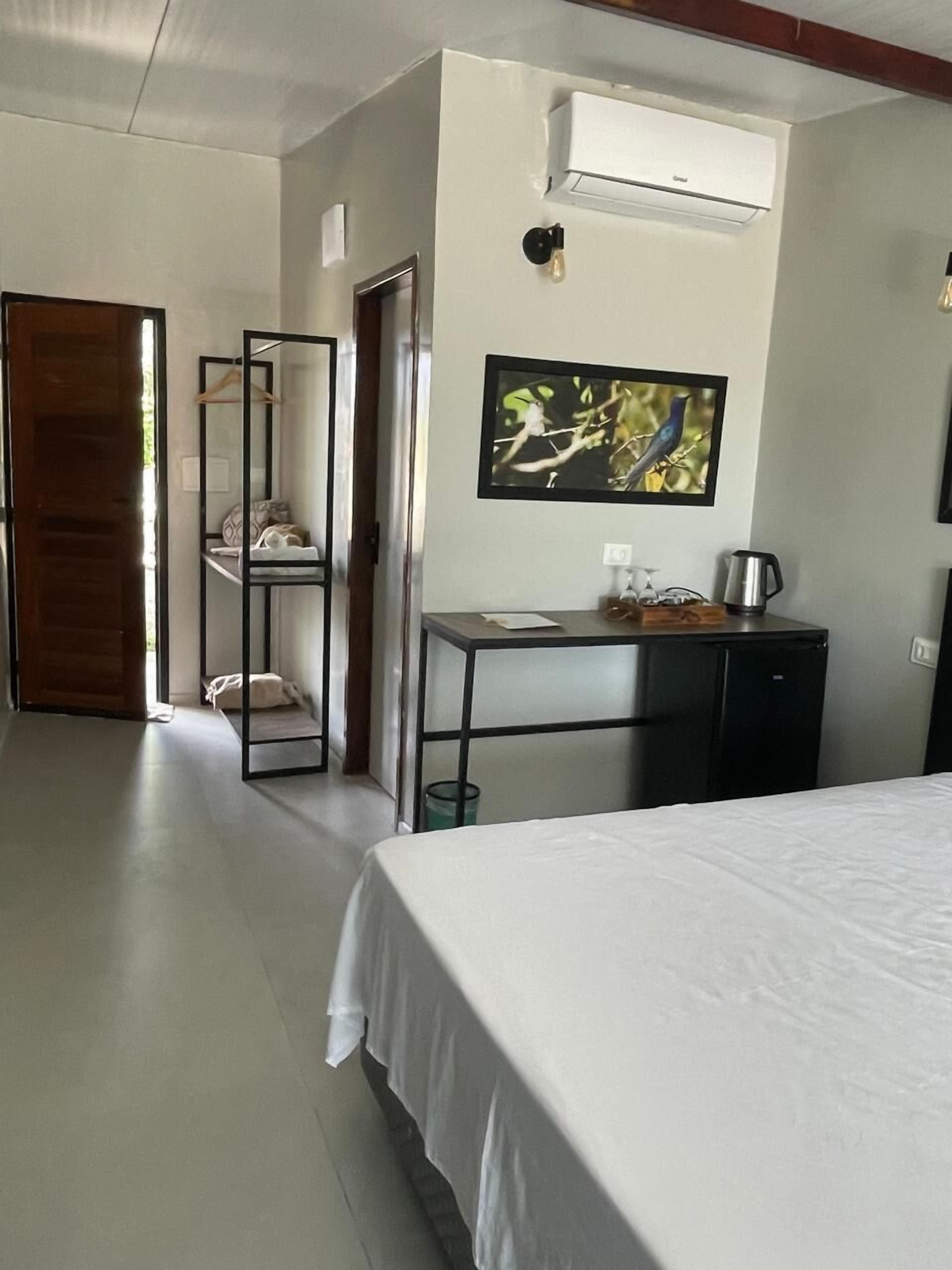 A hotel room with a bed, a desk with a chair, a small kitchenette, and a door leading to another room.