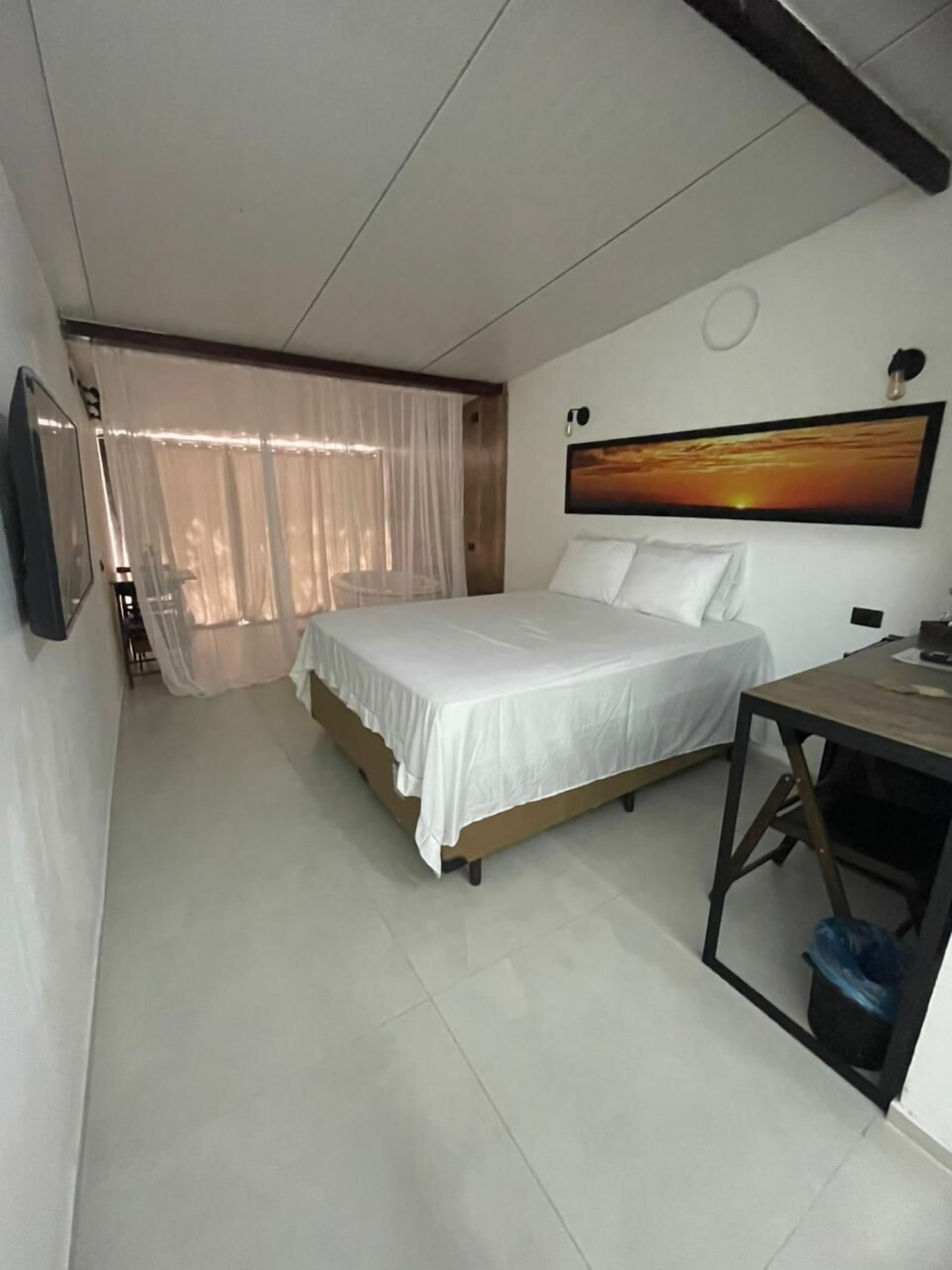 A bedroom with a bed, a desk, a chair, a TV, and a painting on the wall.