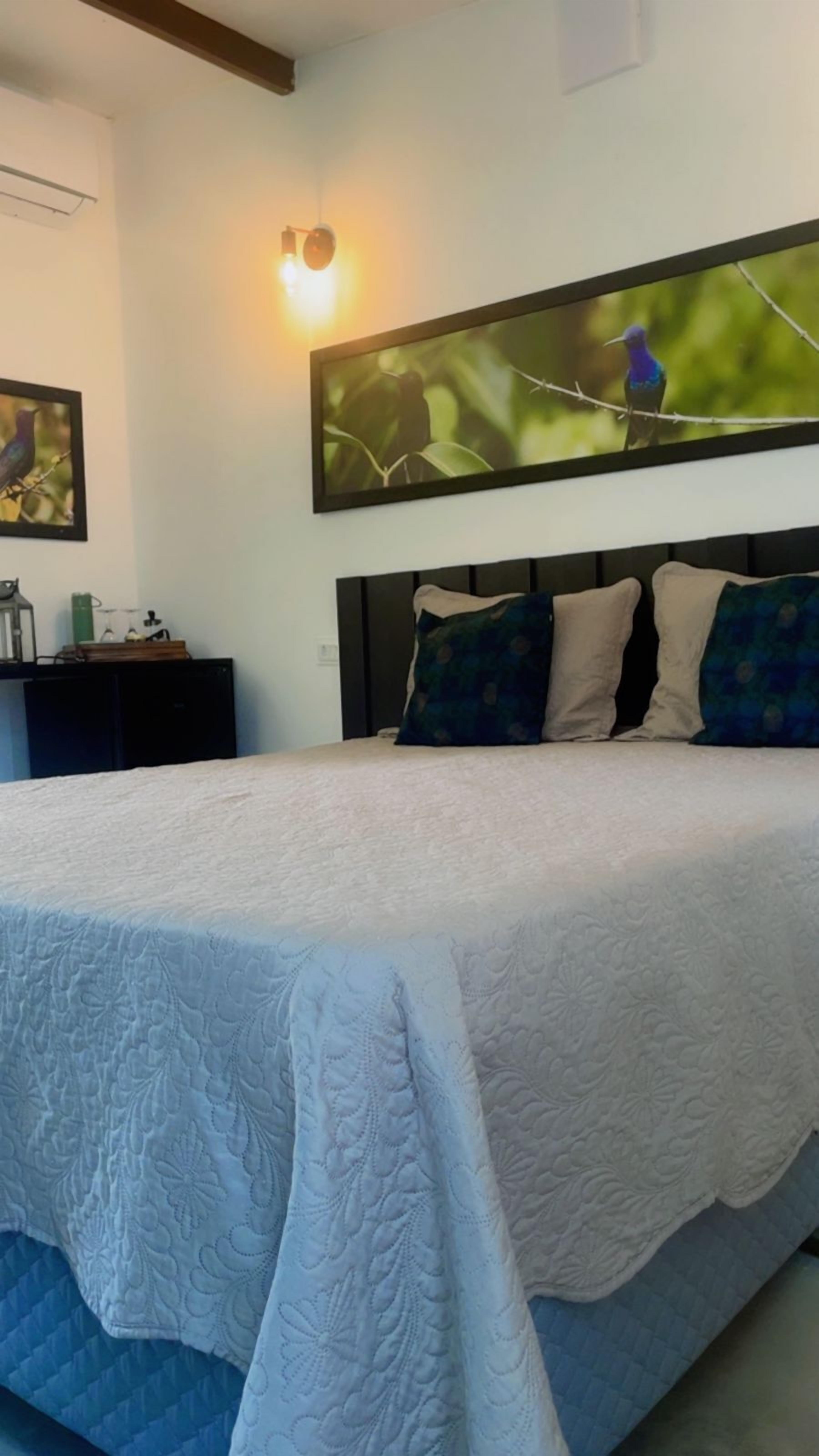 Une chambre à coucher avec un grand lit, une table de chevet, une lampe et une œuvre d’art représentant des oiseaux, accrochée au mur.