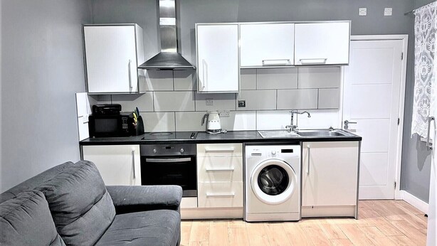 A compact kitchen with white cabinets, a black countertop, a washing machine, and a grey sofa. - Remarkable 1-bed Apartment in High Wycombe (High Wycombe)