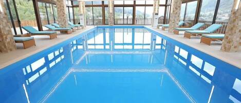 Una piscina interior con agua azul cristalina, rodeada de sillones de descanso y grandes ventanales con vistas al exterior.