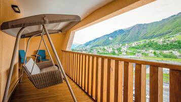 A balcony with a wicker swing chair, wooden railing, and a view of a green valley with buildings.