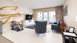 A modern living room with a marble wall, a flat-screen TV, a grey armchair, a sofa, and a dining area with a wooden table and bench. - Apartment 1 of Address Apartments (Gillingham)