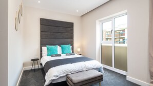 A modern bedroom with a large bed, a bench, a small table, and a view of the outside through a sliding door.