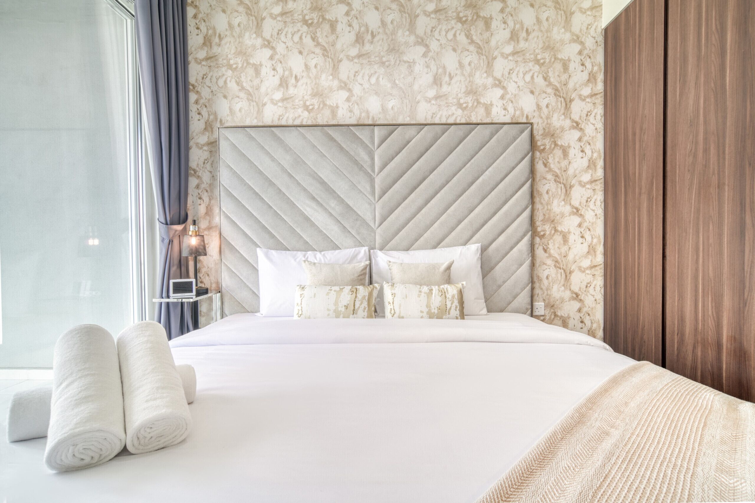 A neatly made bed with a large, textured headboard, a bedside table with a lamp, and a window with curtains.