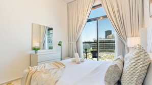 A hotel room with a large bed, a view of the city, a desk with a lamp, and a chair. - Community View 1BR, Just Next to the Park and a Short Walk to the Bus Stop (Dubai)