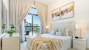 A modern bedroom with a large bed, a nightstand, a bedside table, a potted plant, and a view of the city through large windows. - Community View 1BR, Just Next to the Park and a Short Walk to the Bus Stop (Dubai)
