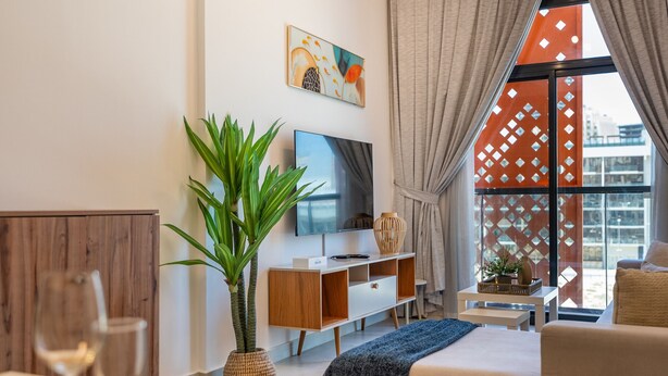 A modern living room with a TV, a potted plant, a sofa, and a dining area with a glass table. - Community View 1BR, Just Next to the Park and a Short Walk to the Bus Stop (Dubai)