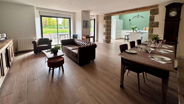 A spacious living area with a sofa, dining table, and chairs. There is a kitchen area with a sink and cabinets, and a large window providing a view of the outdoors.