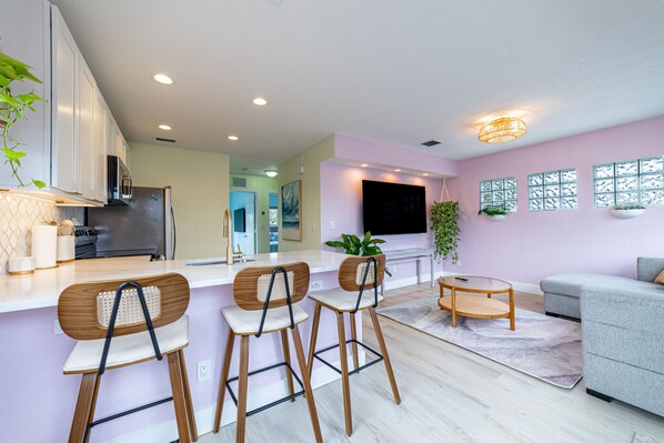 A modern kitchen with white cabinets, a built-in oven, and a refrigerator. There are three wooden bar stools with white cushions. A grey sofa and a small round table are in the living area. - Tropical Treetop Hideaway Just a Short Walk to Beach (Lake Worth Beach)