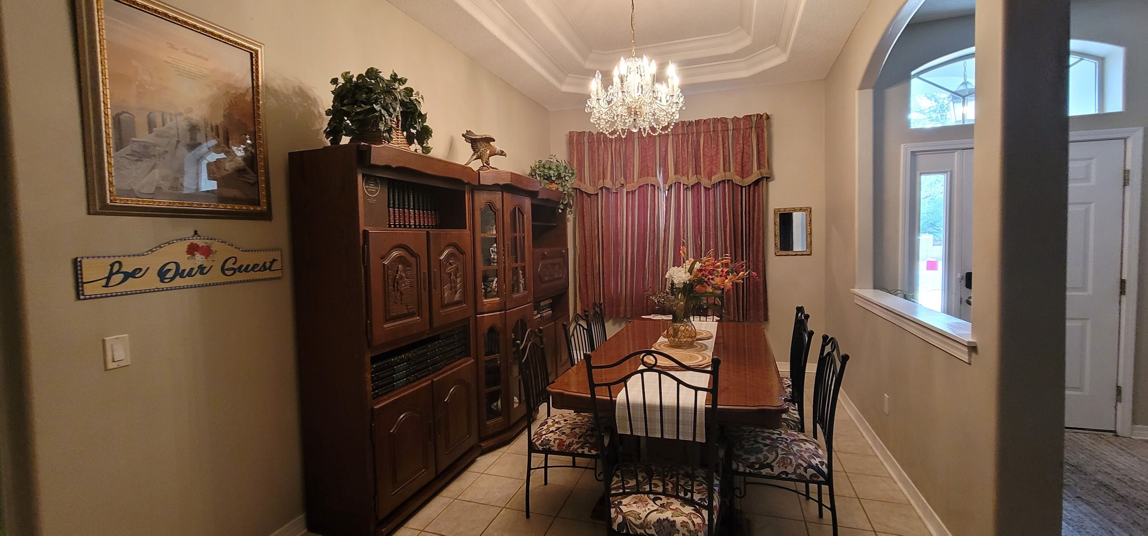 A dining area with a wooden cabinet, a chandelier, and a 'Be Our Guest' sign.